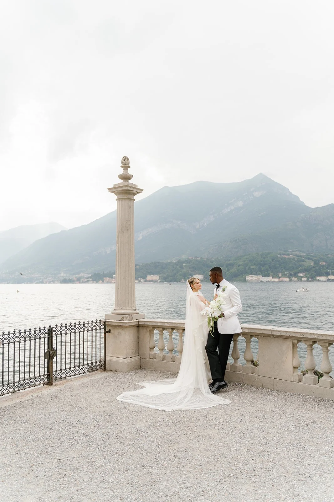 A bride and groom standing on a terrace by a lake with mountains in the background. The bride is in a white wedding gown with a long train, and the groom is in a white tuxedo jacket holding a bouquet of white flowers.