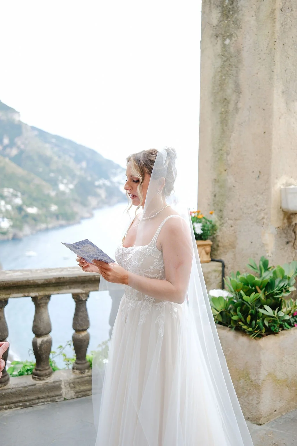 A bride in a white wedding dress and veil reading a letter or card on a balcony with a waterway and mountains in the background.