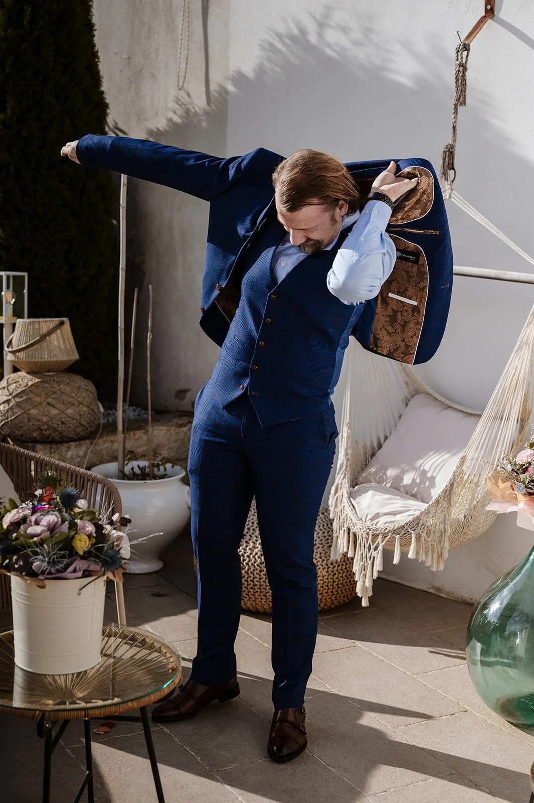 A man in a blue suit putting on a backpack in an outdoor space decorated with flowers, a hammock, and potted plants.