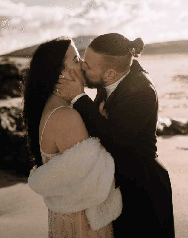 A couple kissing on a beach at sunset, woman wearing a white dress and fur shawl, man in a black tuxedo with a man bun hairstyle.