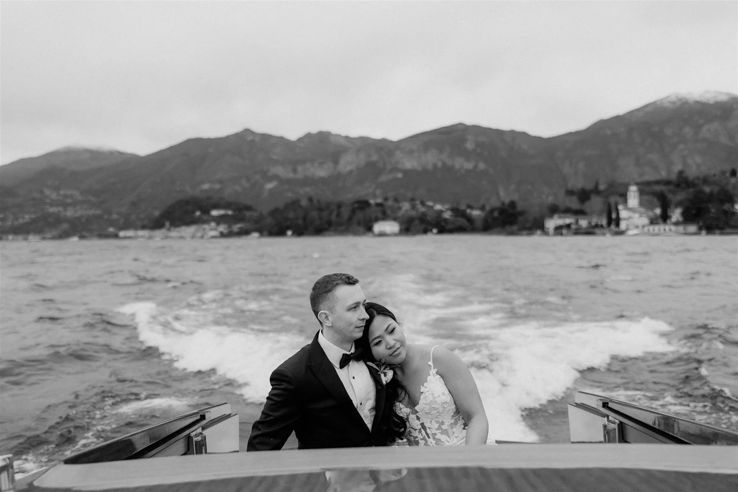 A couple dressed in wedding attire sitting on a boat, embracing each other, with mountains and water in the background.