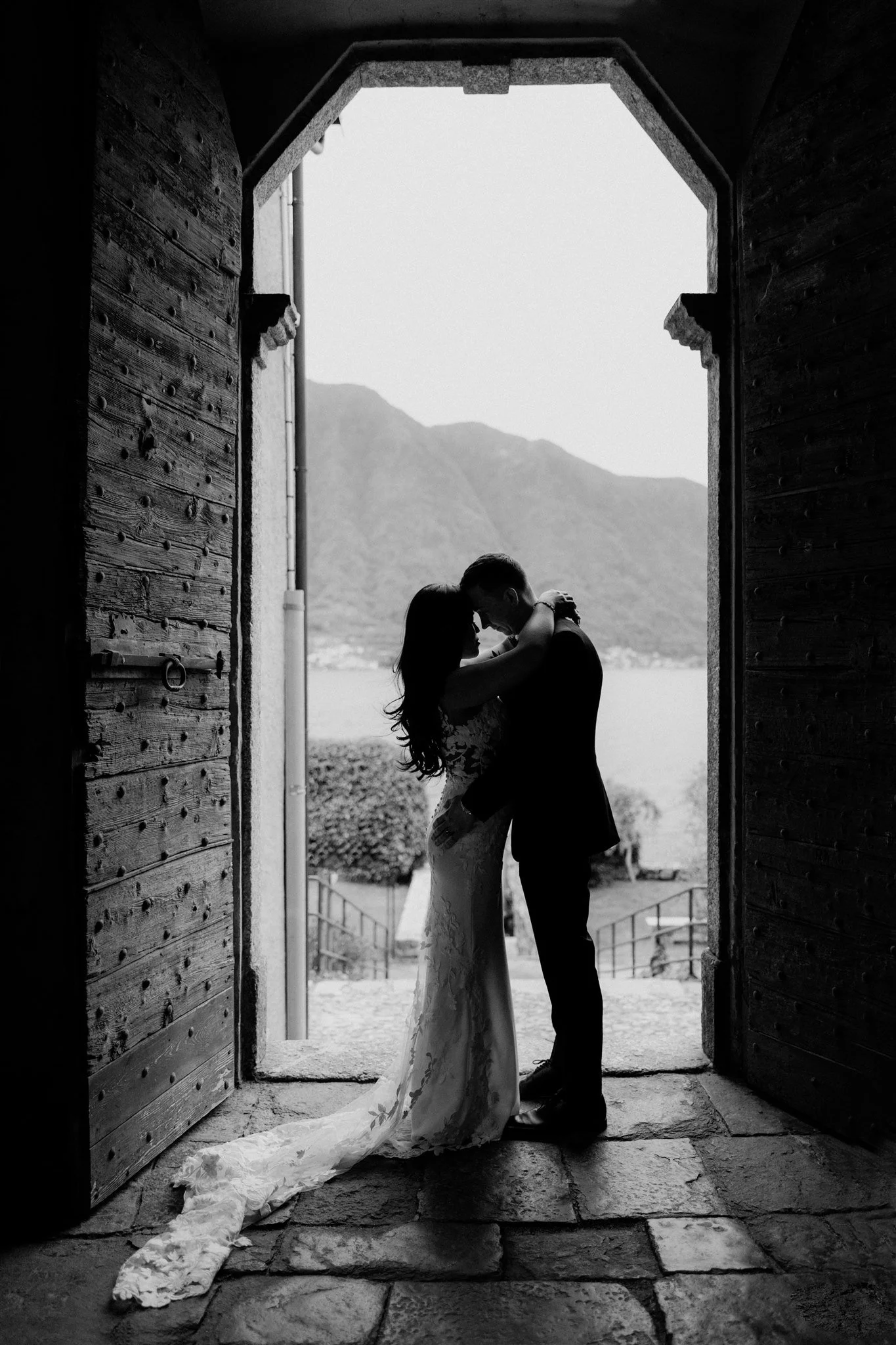A black and white photograph of a couple in wedding attire, standing close and embracing under an open doorway with mountains and a body of water visible in the background.