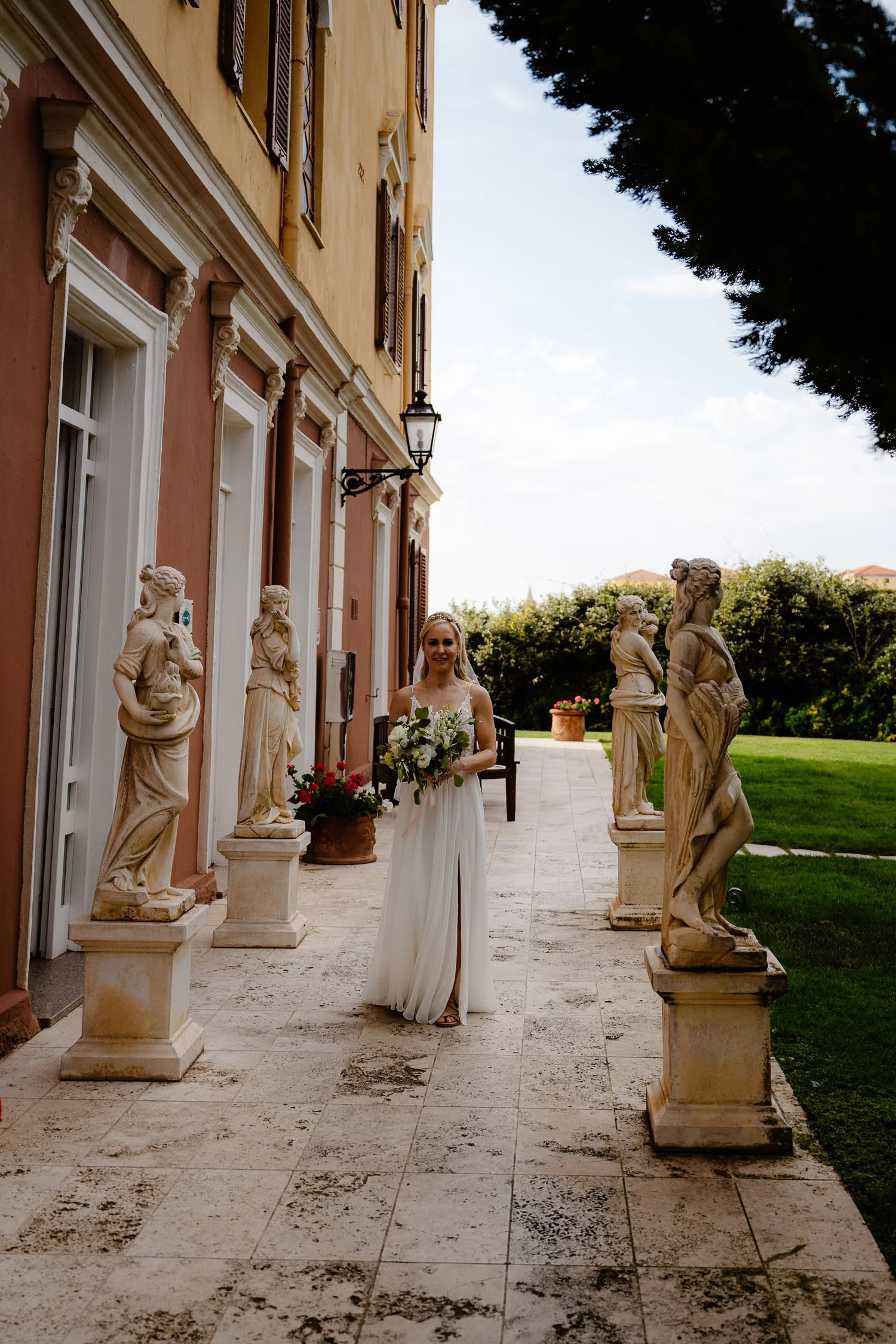 A woman in a white dress holding a bouquet of flowers standing on a tiled patio, surrounded by classical stone statues of women, with a building and greenery in the background.