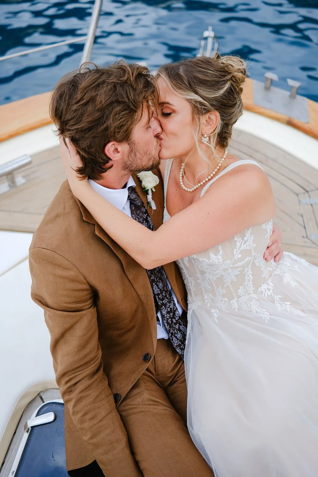 A bride and groom kiss on a boat, with the groom wearing a brown suit and the bride in a white lace wedding gown and pearl jewelry, with water in the background.