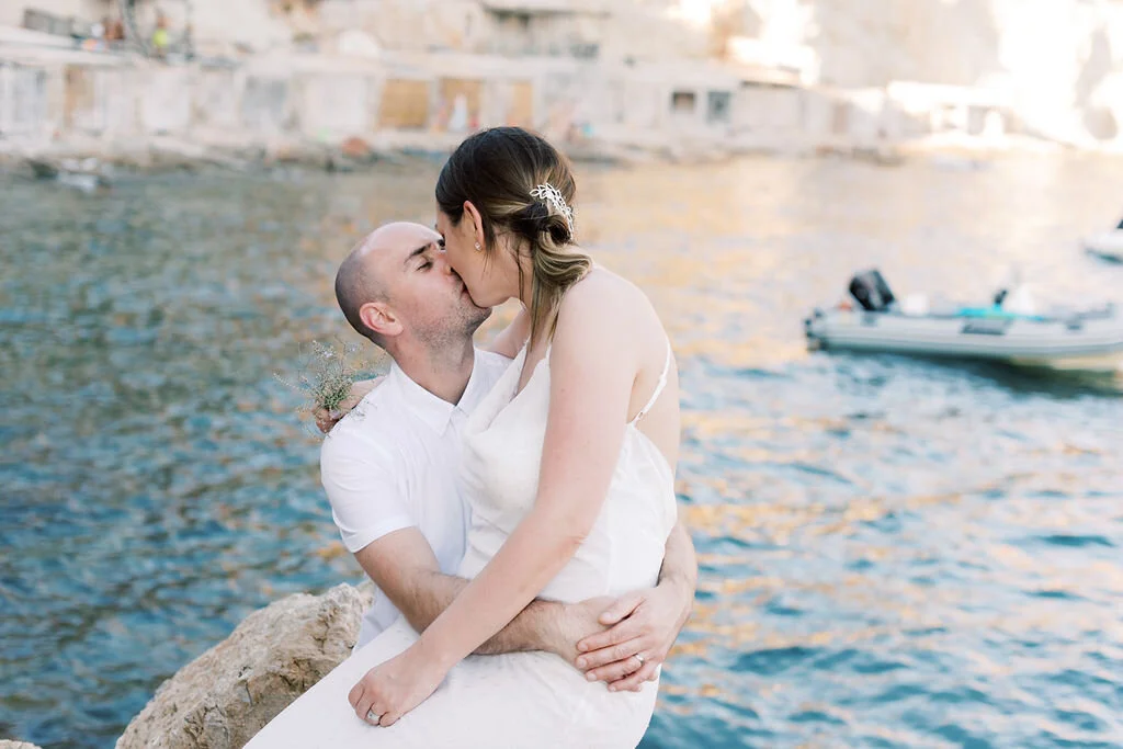 A couple kissing near the water with boats in the background, during sunset.