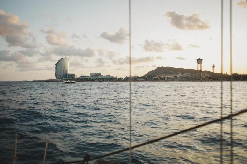 View of Barcelona coastline with W Barcelona hotel, Mount Montjuïc, and water with sailboat at sunset, seen through a boat's window with ropes in the foreground.