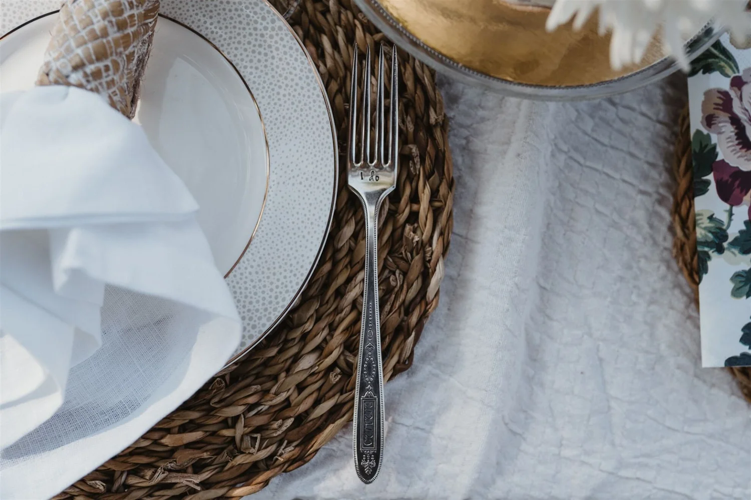 A place setting on a table featuring a white napkin, a decorative white plate with a gold rim, a woven placemat, a fork with intricate design and 'I do' engraved on the handle, a glass with a gold rim, and a floral-patterned cloth underneath the glas
