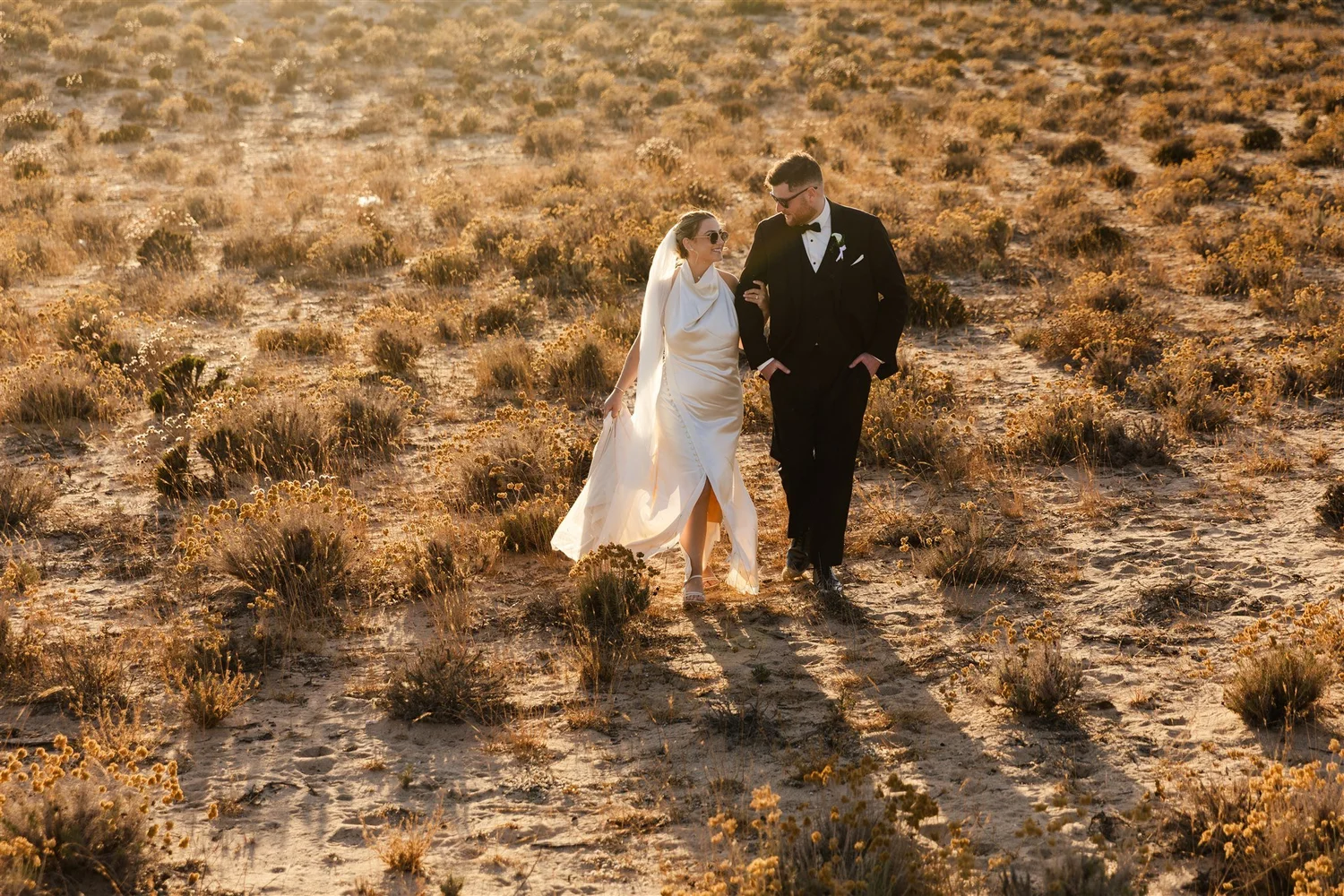 A bride and groom walking together in a desert landscape during sunset, with the bride wearing sunglasses and a white gown, and the groom in a black tuxedo.