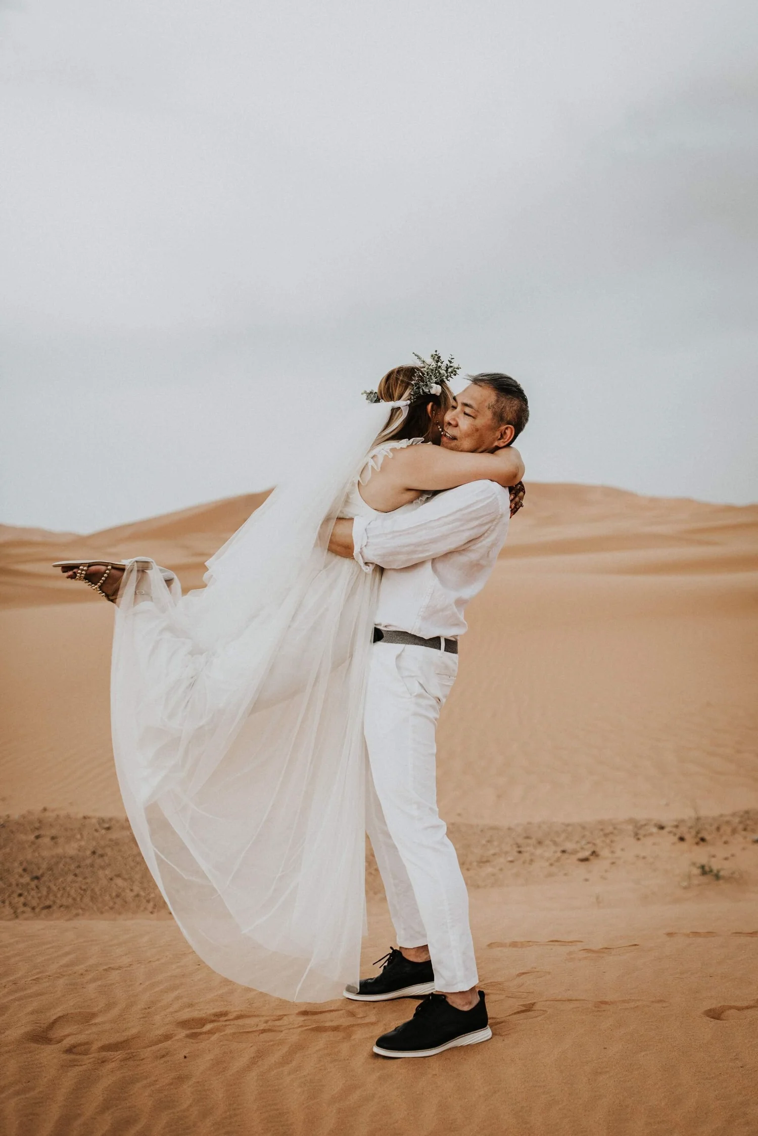 A couple in wedding attire embracing in a desert with sand dunes in the background. The bride is being held in the groom's arms, and they are both smiling, surrounded by an open, natural landscape.