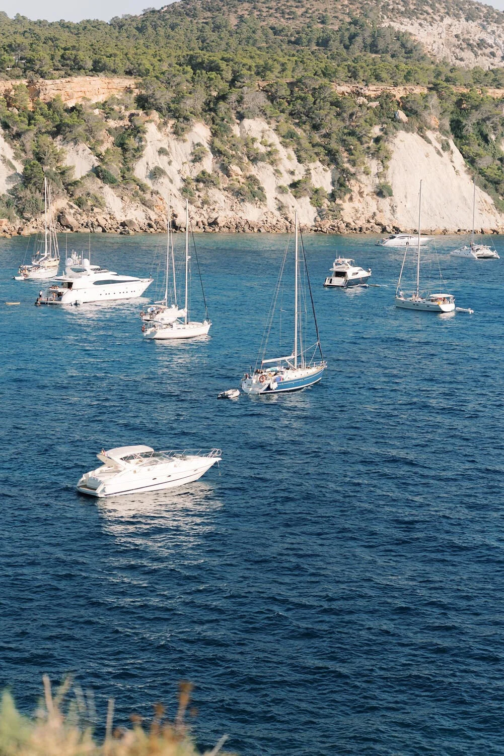 Multiple sailboats and motorboats anchored in a calm, deep blue bay with a rocky and forested hillside in the background.