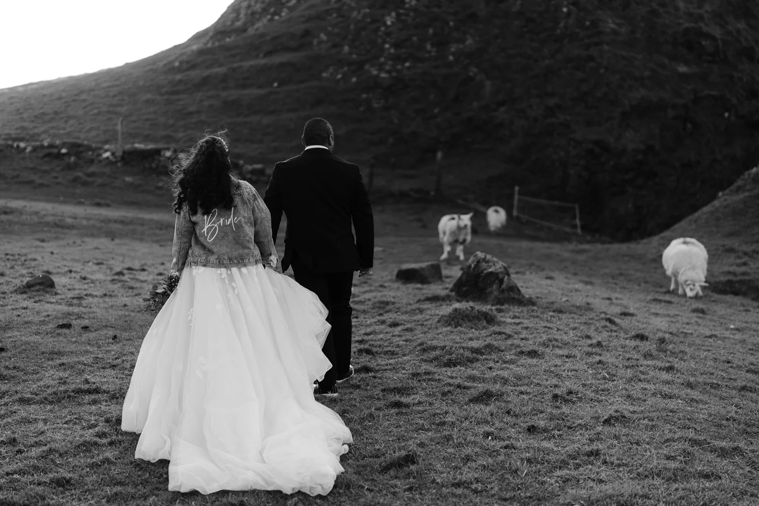 A black and white photo of a bride in a wedding gown and a denim jacket with the word 'Bride' on it, walking hand-in-hand with a groom in a suit in a grassy field with cows and hills in the background.