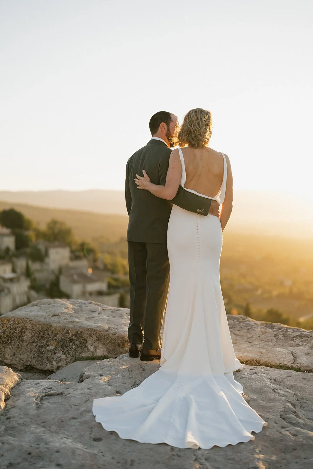 A bride and groom standing close together on a rocky hilltop during sunset, with the bride wearing a white gown with a long train and the groom in a dark suit, overlooking a scenic landscape.