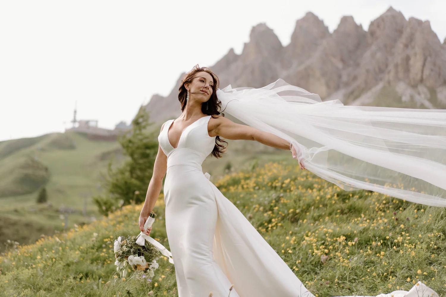 A bride in a white dress holding a bouquet and posing outdoors in a field of wildflowers with mountains in the background.