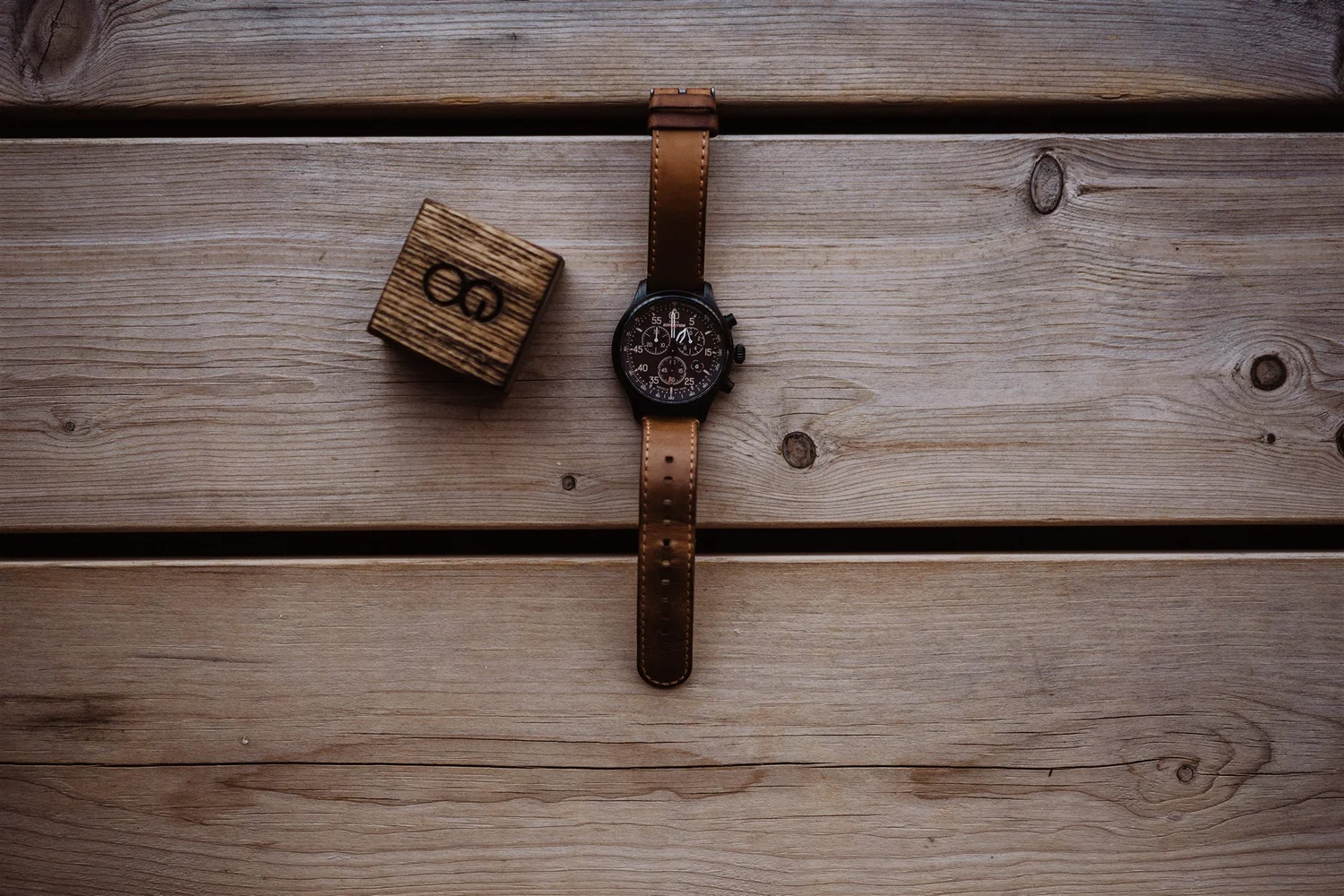 A watch with a black face and tan leather strap placed on a wooden surface along with a wooden block with the number 8 on it.