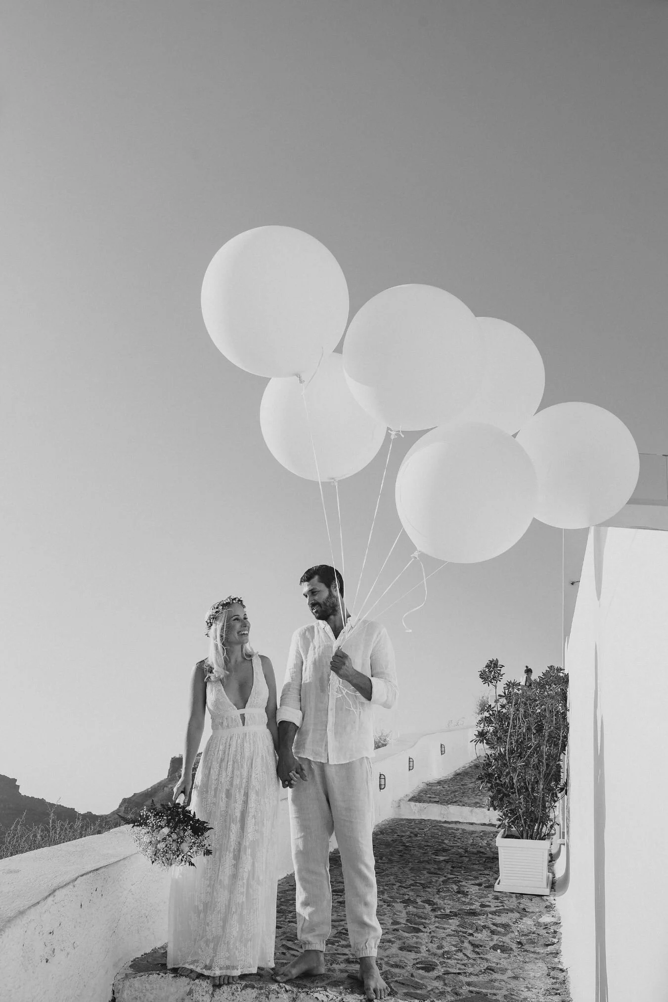 A black and white photo of a bride and groom holding hands and smiling at each other, standing on a stone bench near water. The bride holds a bouquet, and the groom holds a bunch of white balloons. They are outdoors with potted plants and a backdrop