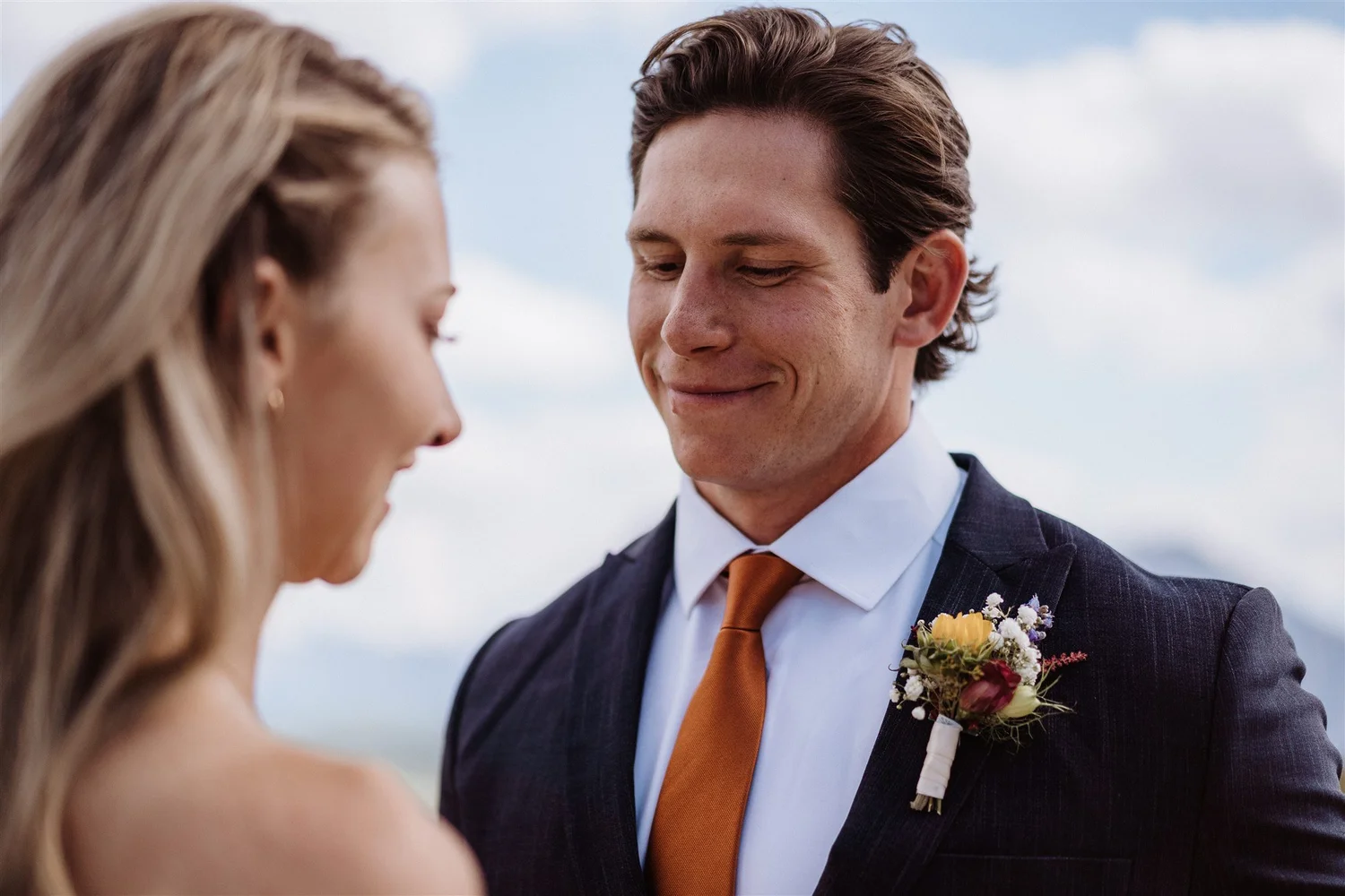 A man and a woman are standing close together outdoors on a cloudy day, facing each other with gentle smiles. The man is wearing a dark suit, white shirt, orange tie, and a boutonnière. The woman has long, light-colored hair, and wears a sleeveless d