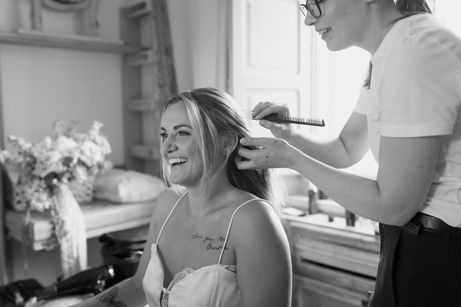 A woman with blonde hair and a tattoo on her chest, smiling while sitting in a chair, as another woman styles her hair with a brush in a bright room.