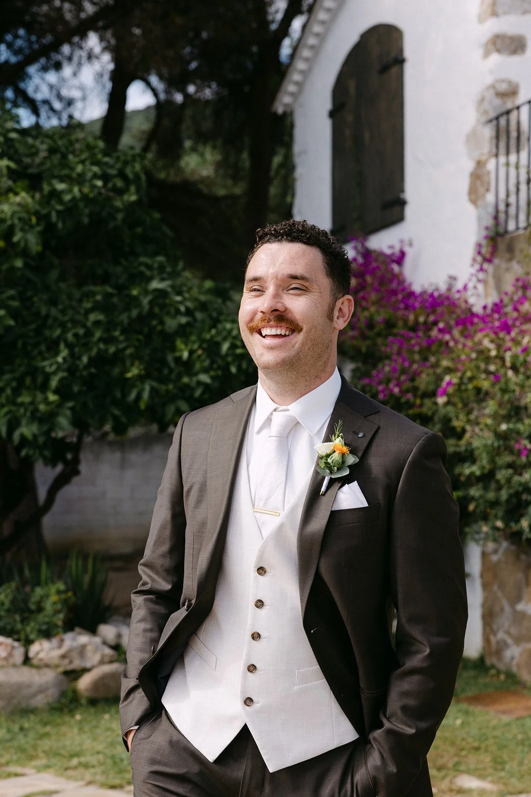 A smiling man dressed in a tuxedo with a boutonniere, standing outdoors with trees, bushes, a white building with black shutters, and pink flowers in the background.