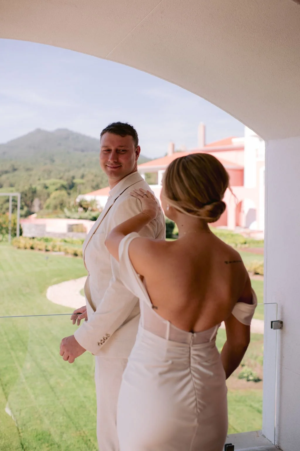 A man and woman stand on a balcony with scenic green hills and a pink building in the background. The man is dressed in a light-colored suit, and the woman wears a silk off-shoulder dress. She touches his shoulder, and he looks back smiling.