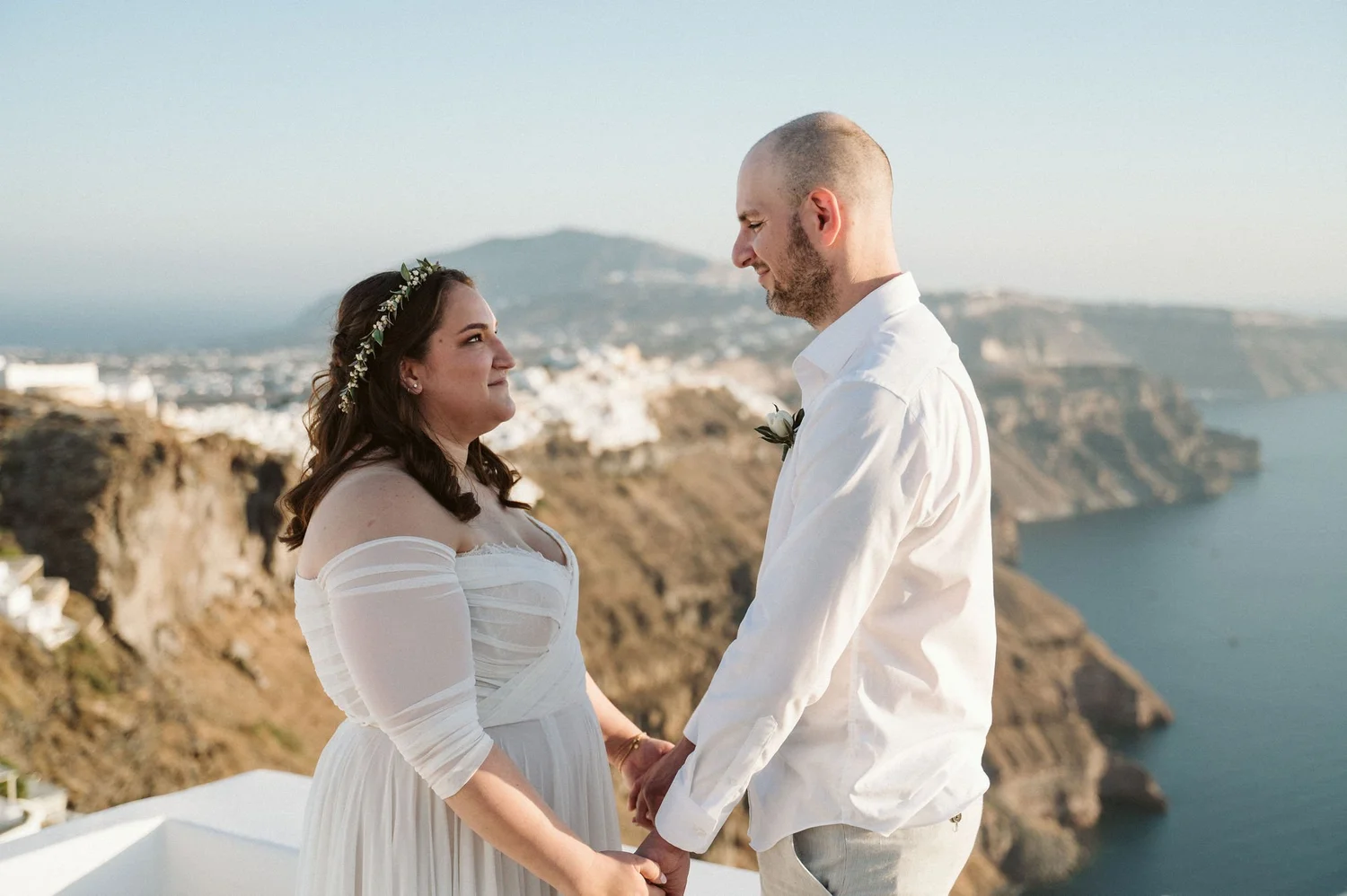A bride and groom holding hands and facing each other on a cliffside overlooking the ocean at sunset, with white buildings and a mountain in the background.