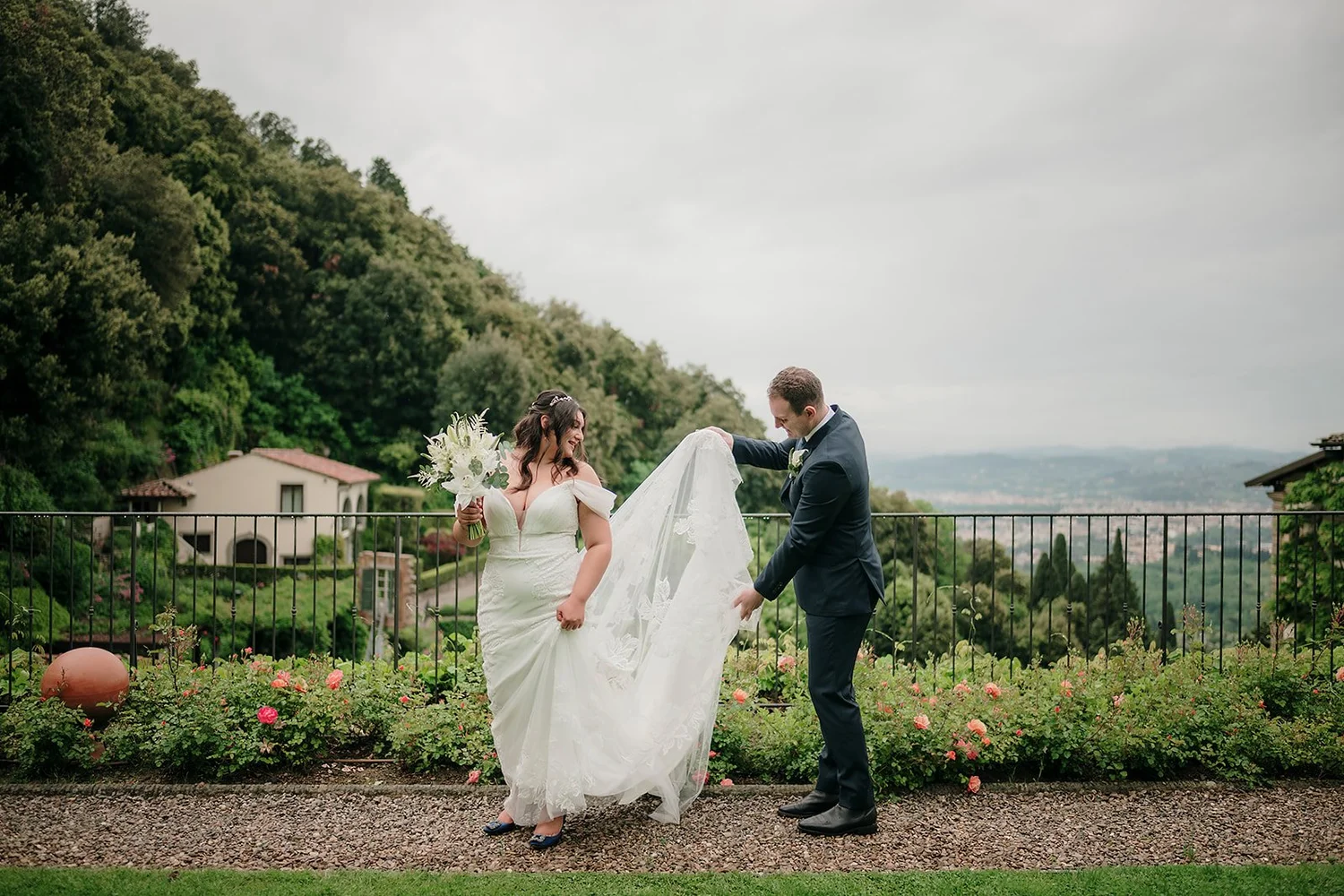 A bride in a white wedding gown holding a bouquet of flowers and smiling at a groom in a dark suit. The groom is lifting the bride's veil outdoors with a scenic hilly landscape in the background.