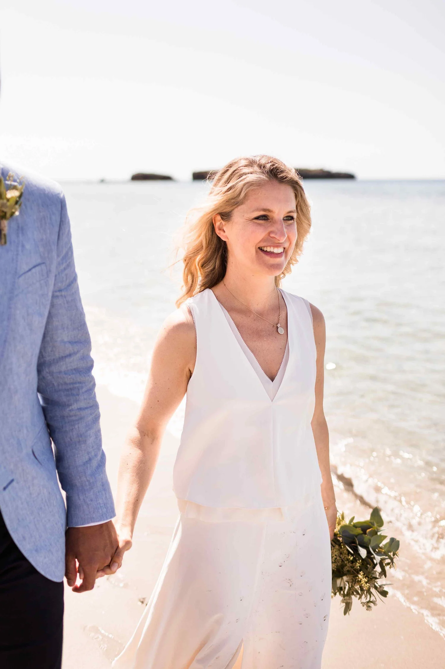 A woman in a white dress holding hands with a man in a light blue suit, standing on a beach with the ocean in the background, during daytime.