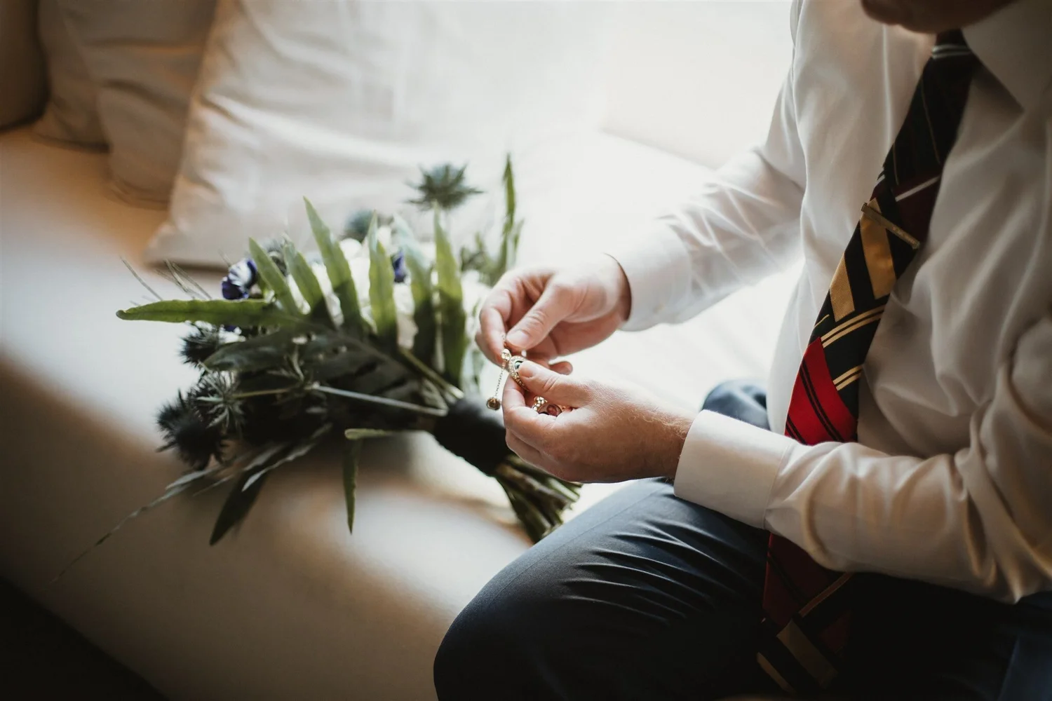 Person in a white shirt, striped tie, and gray trousers sitting on a windowsill, holding a silver necklace with a pendant amidst a bouquet of flowers with blue and white petals.