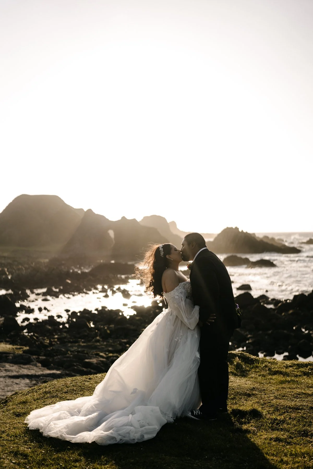 A bride and groom kissing on a rocky shoreline at sunset, with the ocean and cliffs in the background.