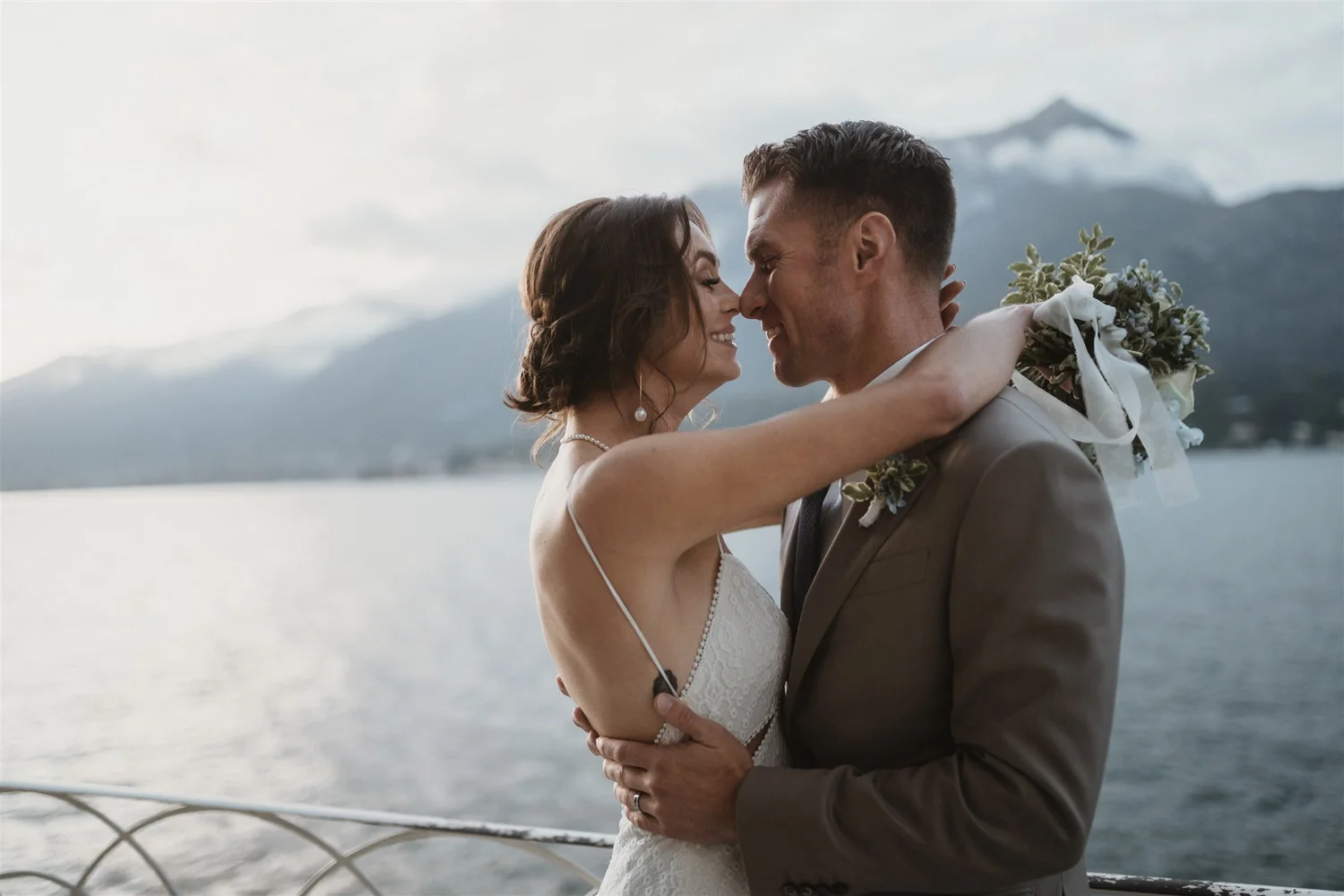 A bride and groom embrace and smile with their noses touching, standing outdoors near a body of water with mountains and fog in the background.