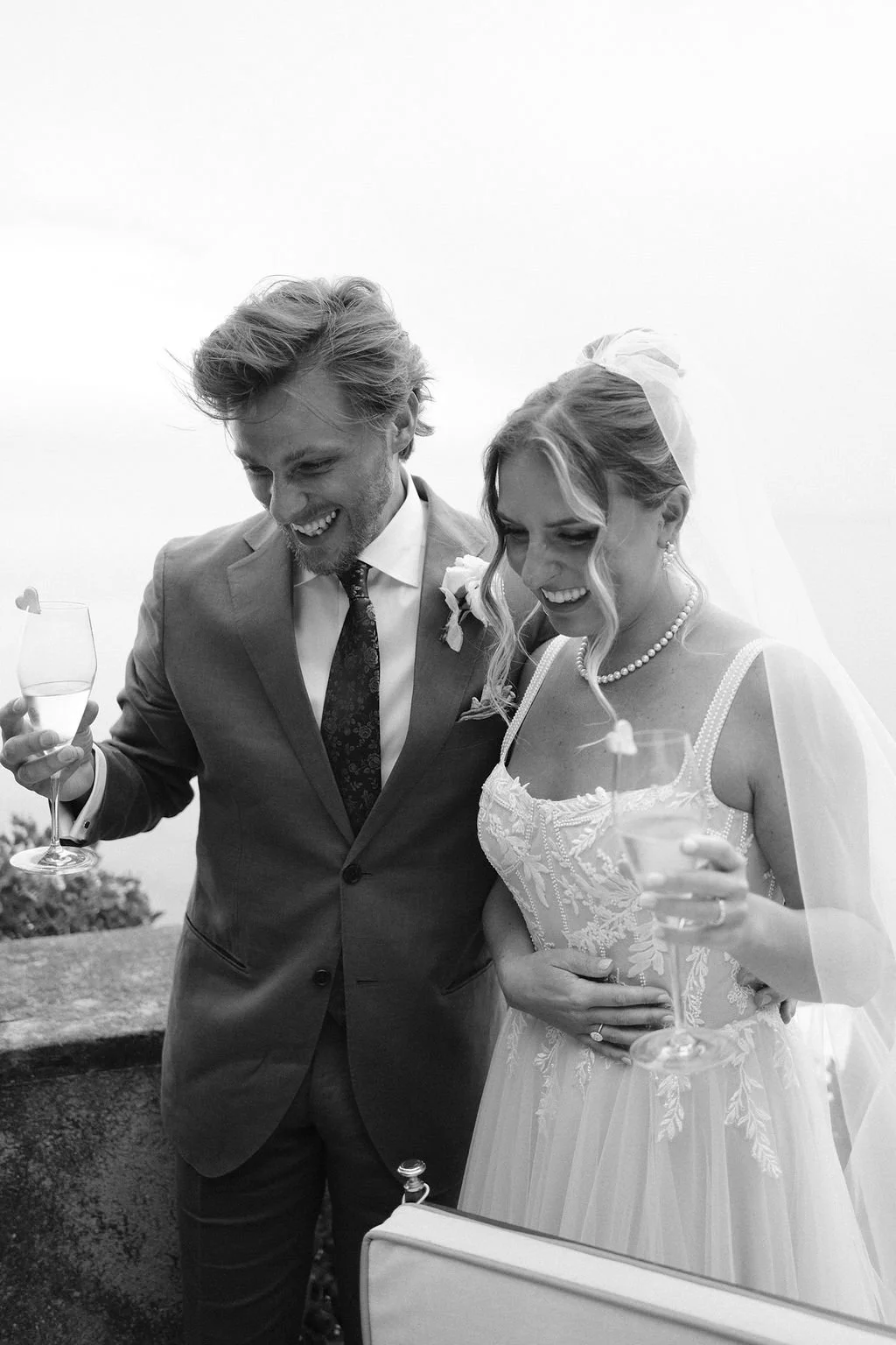 A black-and-white photo of a happy wedding couple, the groom with tousled hair in a suit and the bride with a veil and pearl necklace, holding glasses of champagne and smiling.