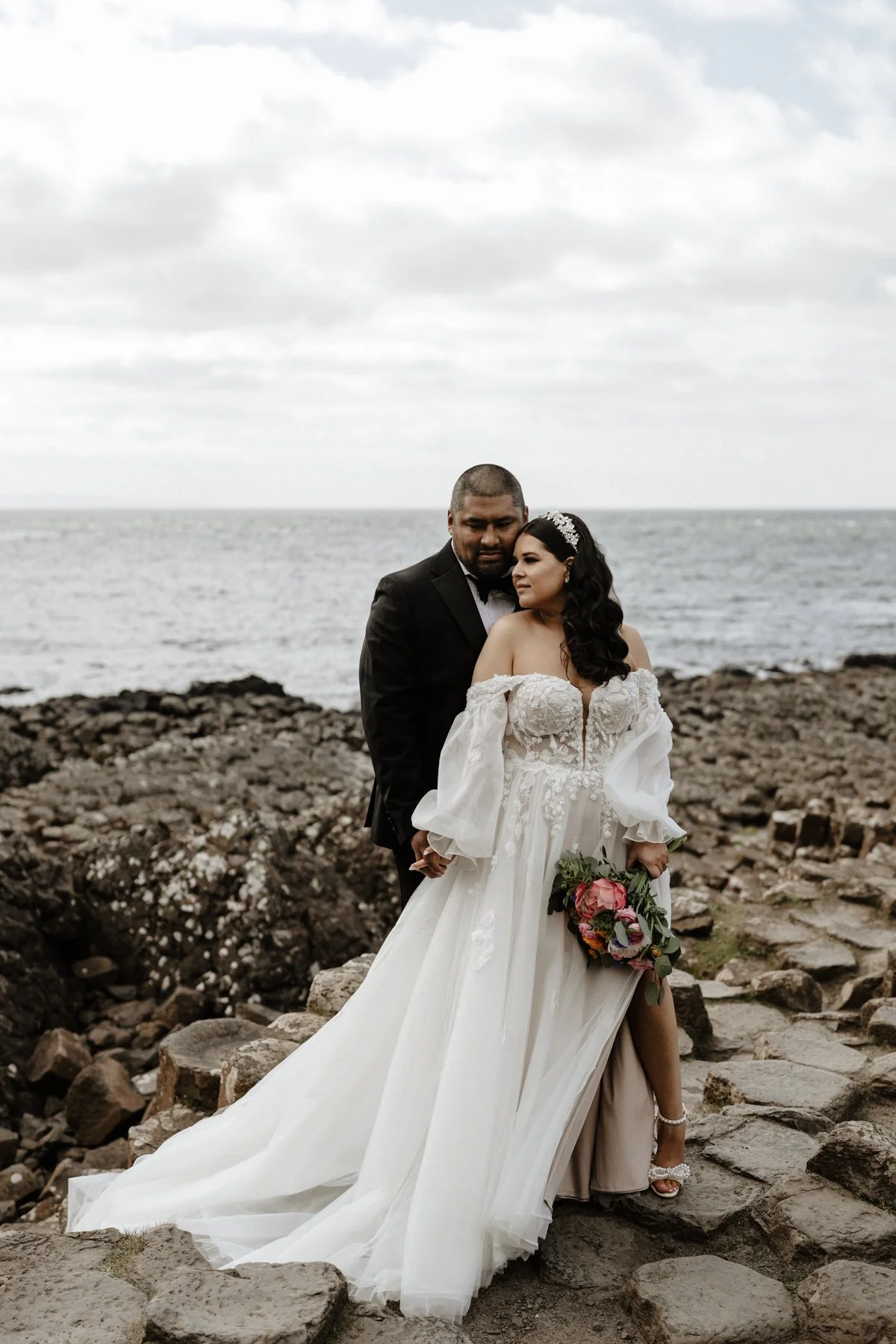 A newlywed couple dressed in wedding attire standing on rocky terrain by the ocean, with cloudy sky overhead.