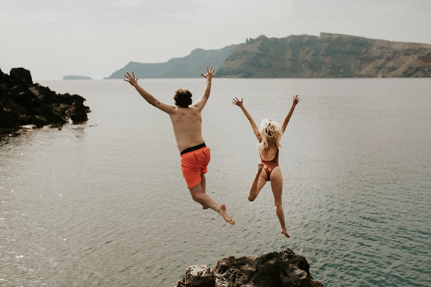 A man and woman jumping into the water at a rocky seaside with mountainous background, wearing swimwear.