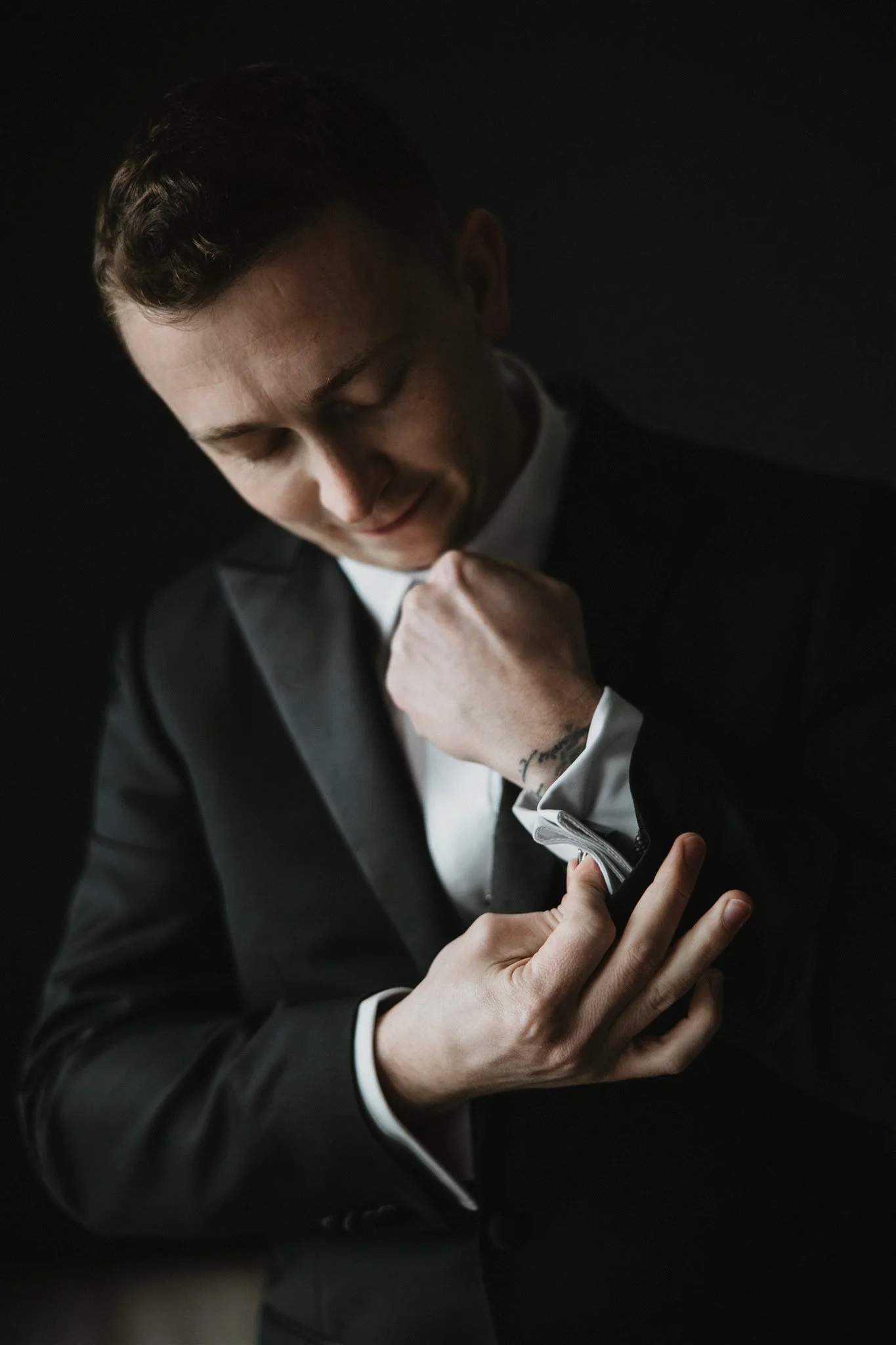 A man in a black tuxedo adjusting his white shirt and tie, with a confident smile, against a dark background.