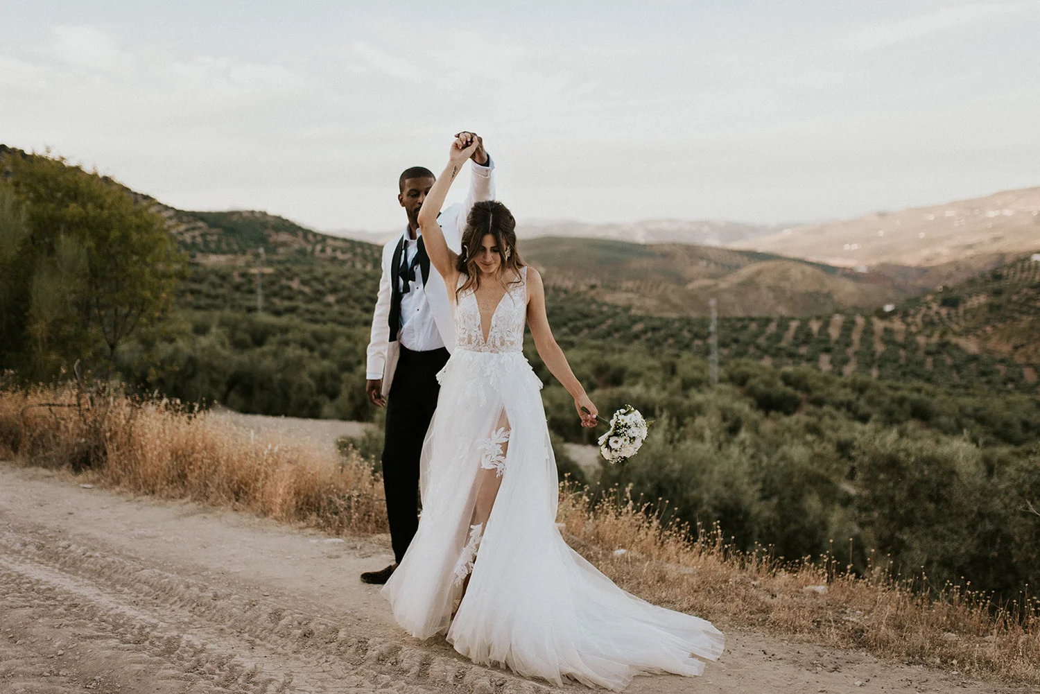 A bride and groom walking outdoors on a dirt path in a rural landscape with rolling hills in the background. The bride is wearing a white wedding dress and holding a bouquet of flowers, while the groom is dressed in a white blazer, black shirt, and b
