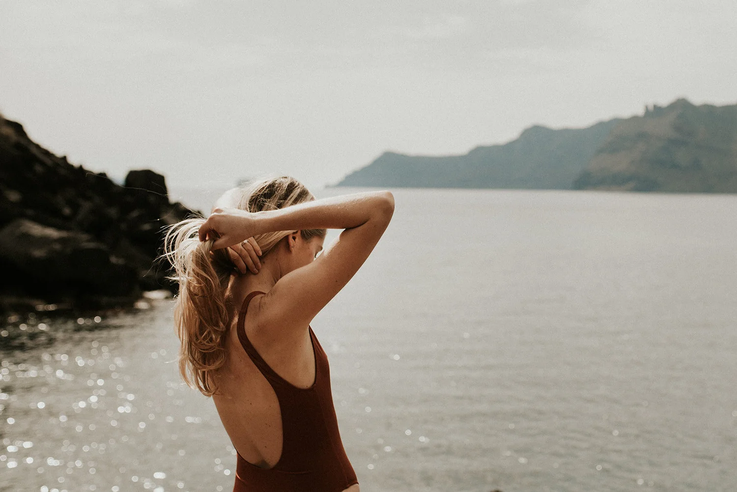 A woman with blonde hair tying her hair at the beach with mountains in the background.