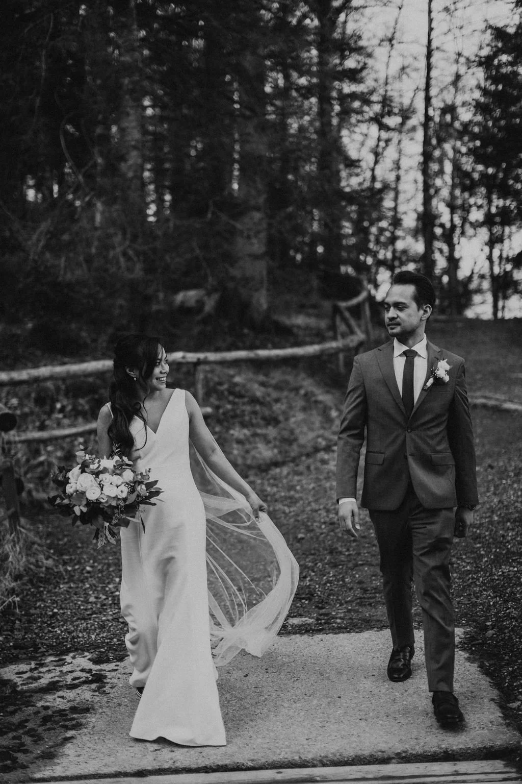 A bride and groom walking through a wooded outdoor area, with the bride smiling and holding a bouquet of flowers, and the groom looking at her, dressed in wedding attire.