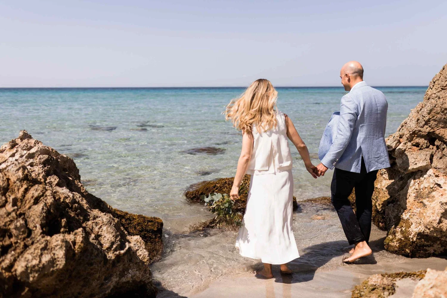 A couple dressed in wedding attire holding hands and walking on a rocky beach near the ocean.
