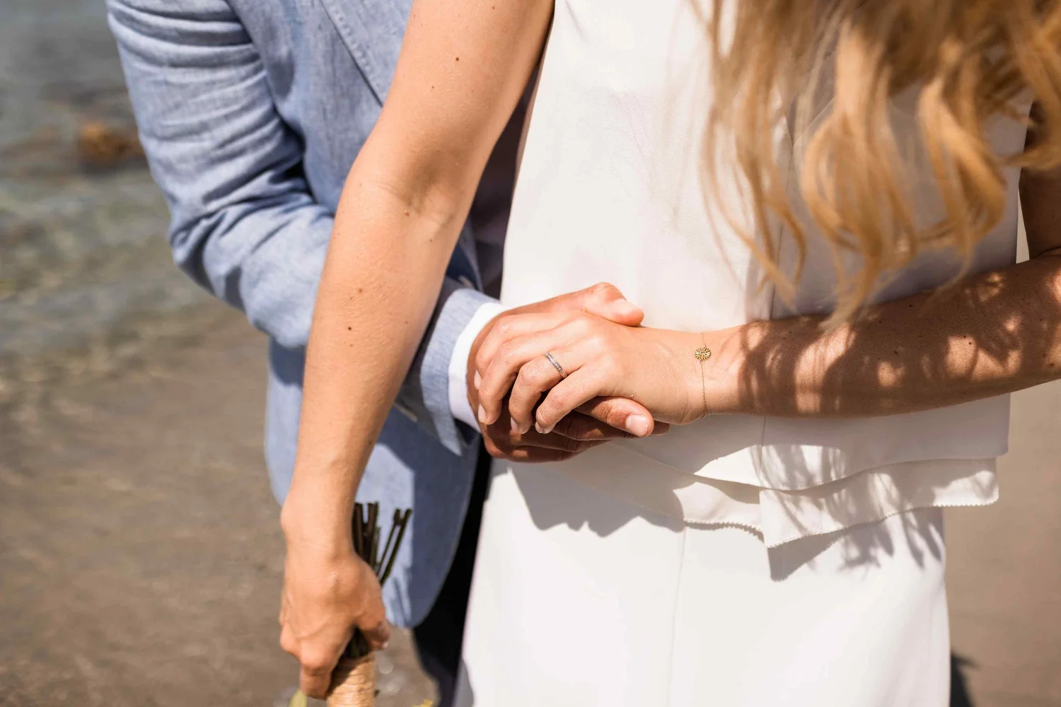 A close-up of a couple holding hands, with the woman's hand displaying a wedding ring and a delicate bracelet, and the man's hand holding her hand from below. The background suggests they are outdoors near a sandy or beach area.