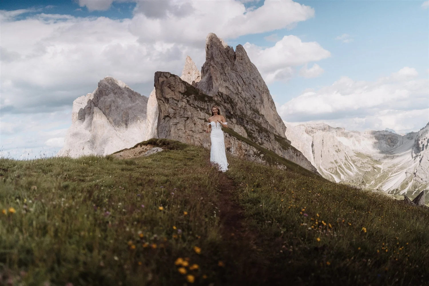 A woman in a white dress standing on a grassy hillside with large rocky mountains and a partly cloudy sky in the background.