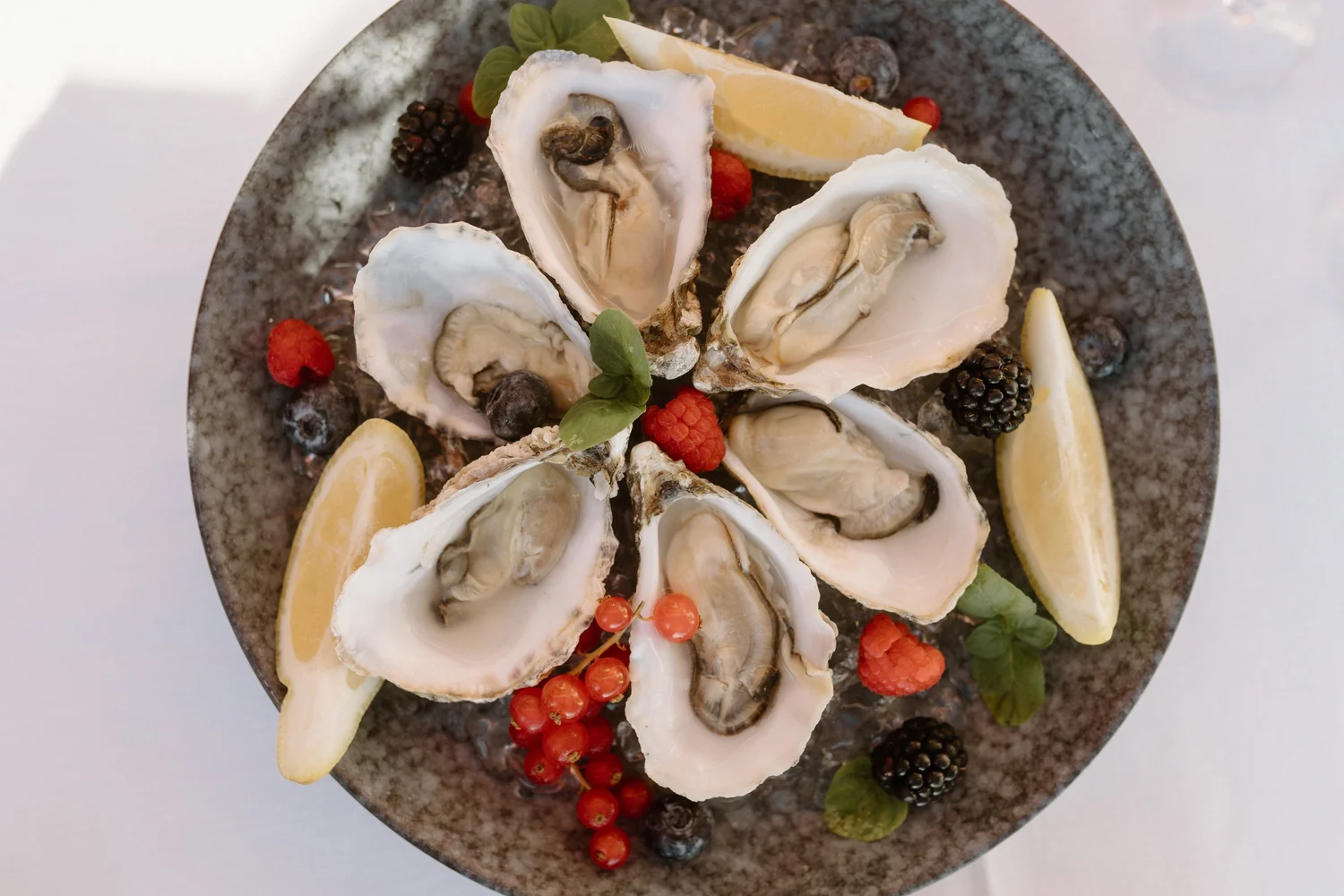 A plate of fresh oysters on a dark ceramic dish, garnished with lemon wedges, red currants, blackberries, raspberries, and green herbs.