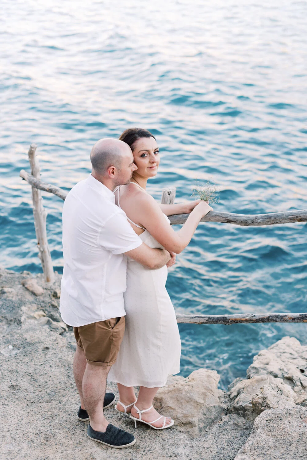 A man and woman standing on rocks near a wooden railing by the water, with the man embracing the woman from behind, they are looking at the camera, the woman is holding a bunch of flowers or greenery, and the water is calm with light ripples.