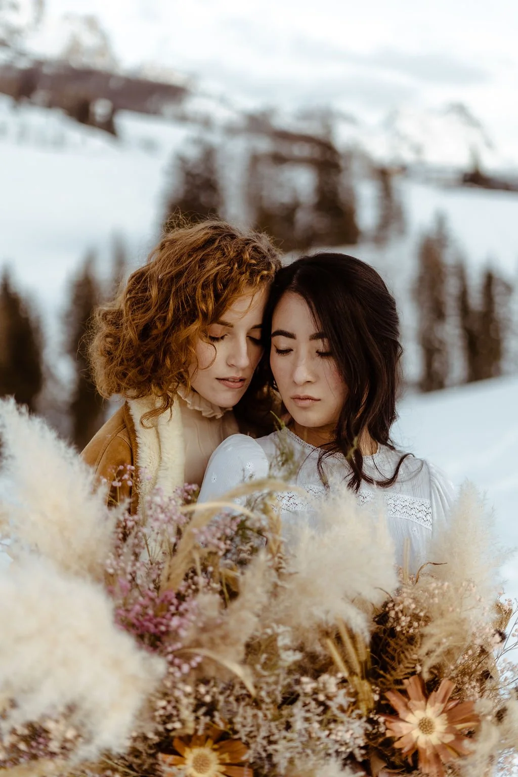 Two women with their eyes closed, standing closely together amidst a field of dried flowers, with snow-covered trees and a ski lift in the background.