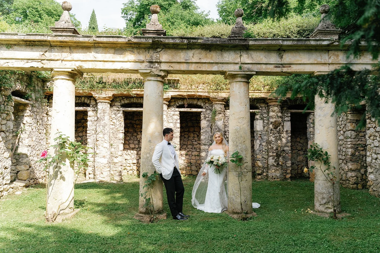 A bride and groom standing in an ancient stone and columns structure surrounded by greenery. The groom is wearing a white tuxedo jacket and black pants, while the bride is holding a bouquet of flowers and wearing a white wedding dress.
