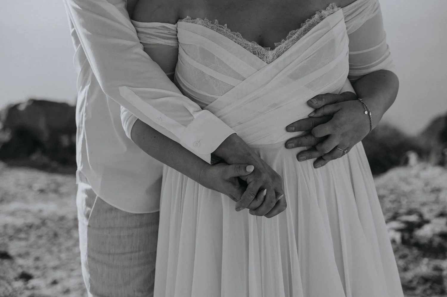 A close-up black and white photo of a person wearing a wedding dress, holding hands with someone in an outdoor setting.