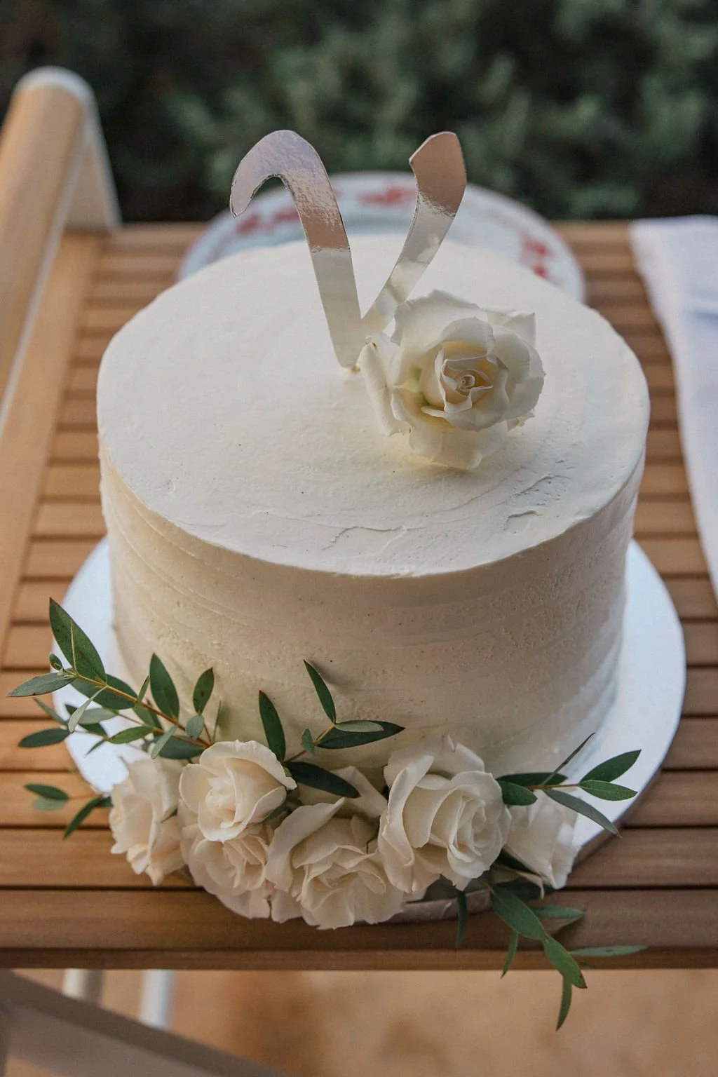 A white frosted cake decorated with white roses and green leaves, topped with a silver heart-shaped cake topper, on a wooden surface.