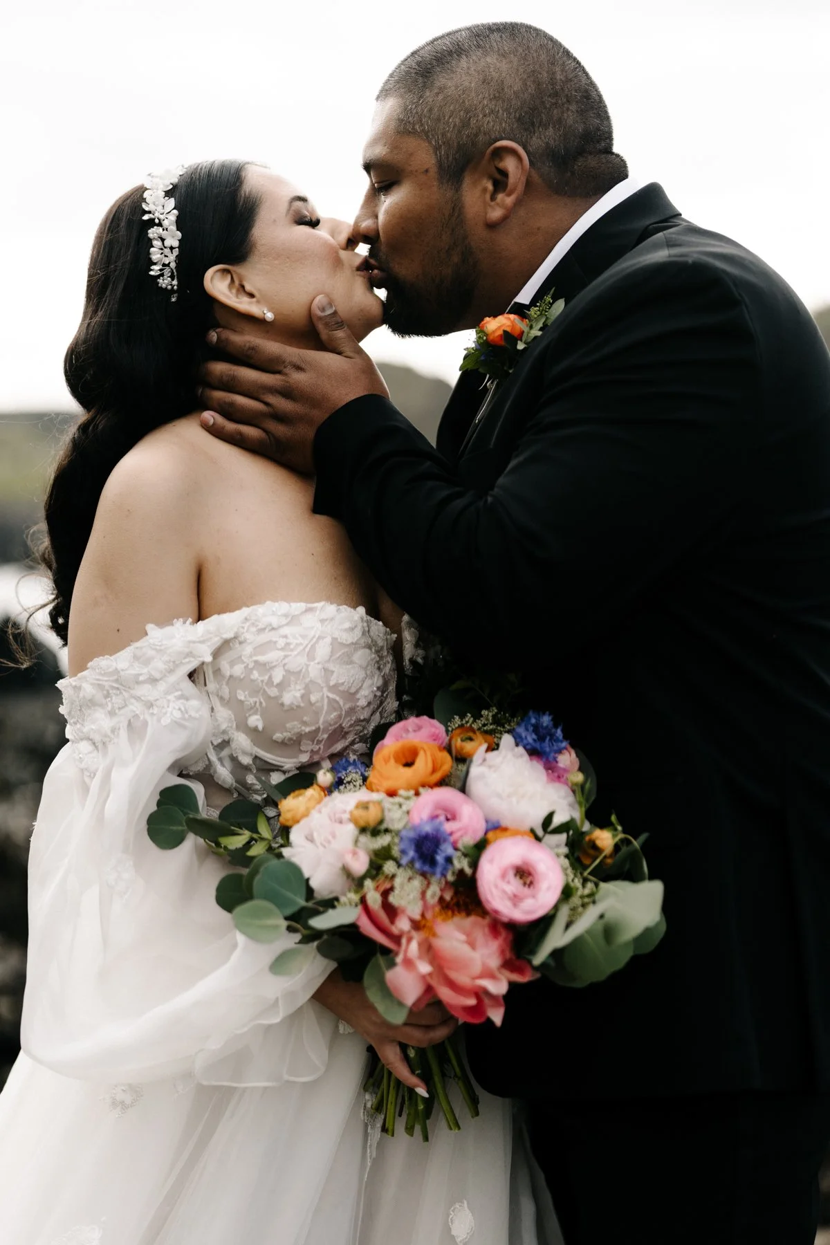 Bride and groom sharing a kiss, holding a colorful bouquet of flowers. The bride is wearing an off-the-shoulder lace wedding dress and a floral headpiece. The groom is dressed in a black tuxedo with a boutonniere.
