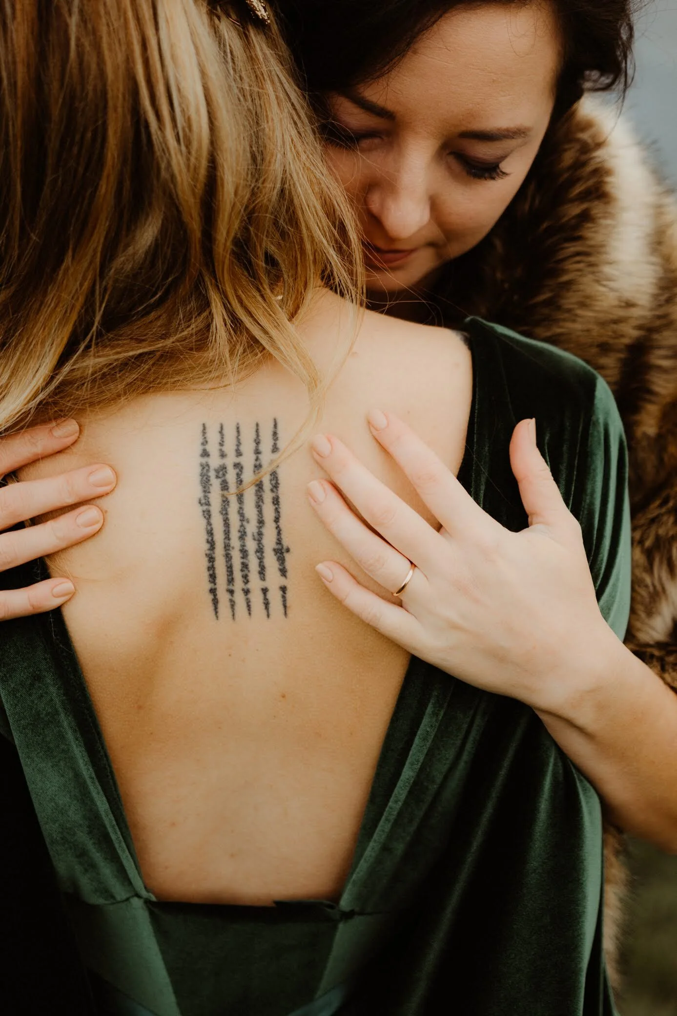 A woman with dark hair embraces another person, showing a tattoo of lines of text on her shoulder. The woman wears a green velvet dress and has her hands resting on her skin.