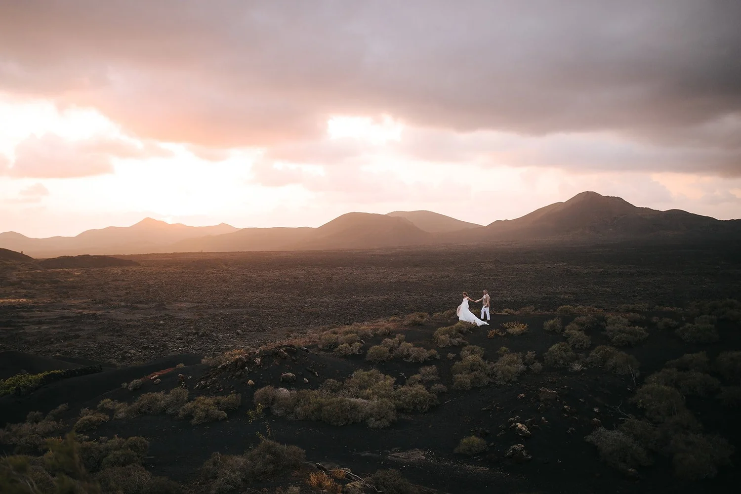 A bride and groom holding hands and walking through a volcanic landscape at sunset, with mountains in the background and dramatic clouds overhead.