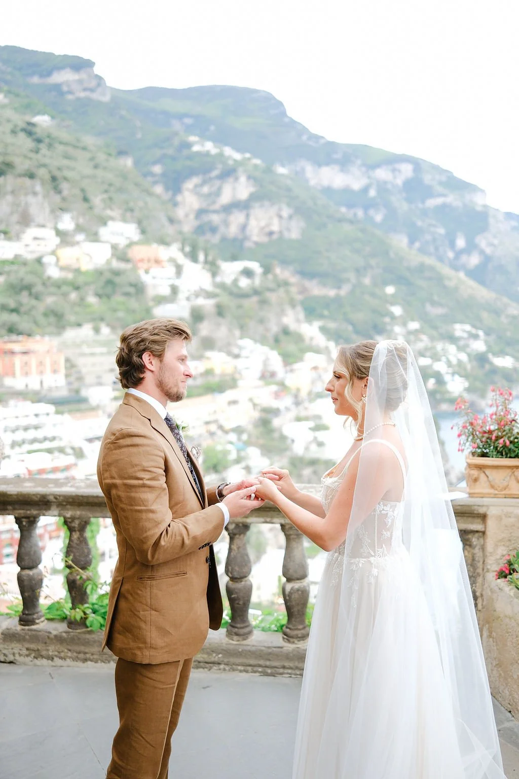 A bride and groom exchange wedding vows on a balcony overlooking a hillside town, with mountains in the background.