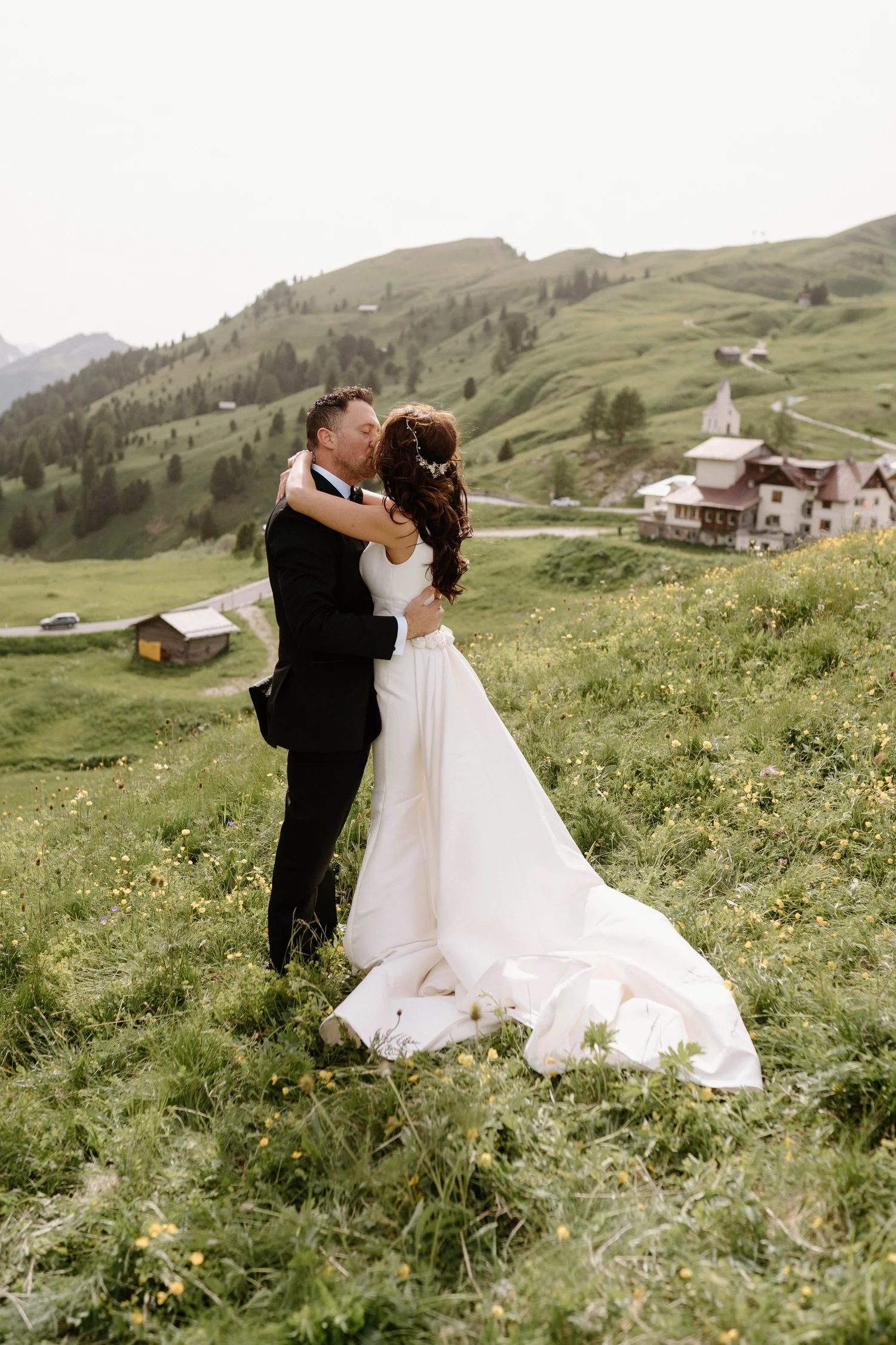A bride and groom sharing a kiss outdoors in a green meadow with hills and small houses in the background.